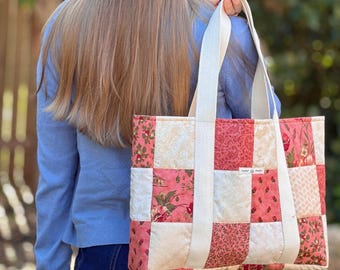 May include: A patchwork tote bag in pink, cream, and red hues. The bag has a floral and solid square design, with cream-colored straps. The bag is carried over the shoulder. The bag is made of quilted fabric.