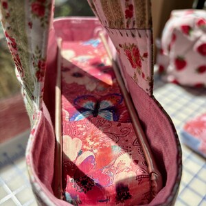 May include: A handmade pink fabric tote bag with a floral and butterfly pattern. The interior features a pink lining and a decorative butterfly design. The bag has a rectangular shape and a handle.