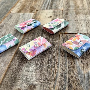 May include: Several small, rectangular cardholders with floral patterns in pink, yellow, and blue are arranged on a wooden surface. Each cardholder has a silver snap closure. The cardholders have a rounded shape.
