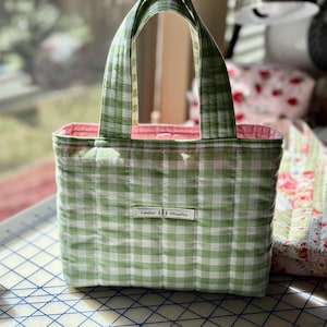 May include: A small, quilted tote bag with a green and white gingham pattern. The bag has a pink interior and matching green handles. A small white tag with the text "Cedar Studios" is sewn on the front.