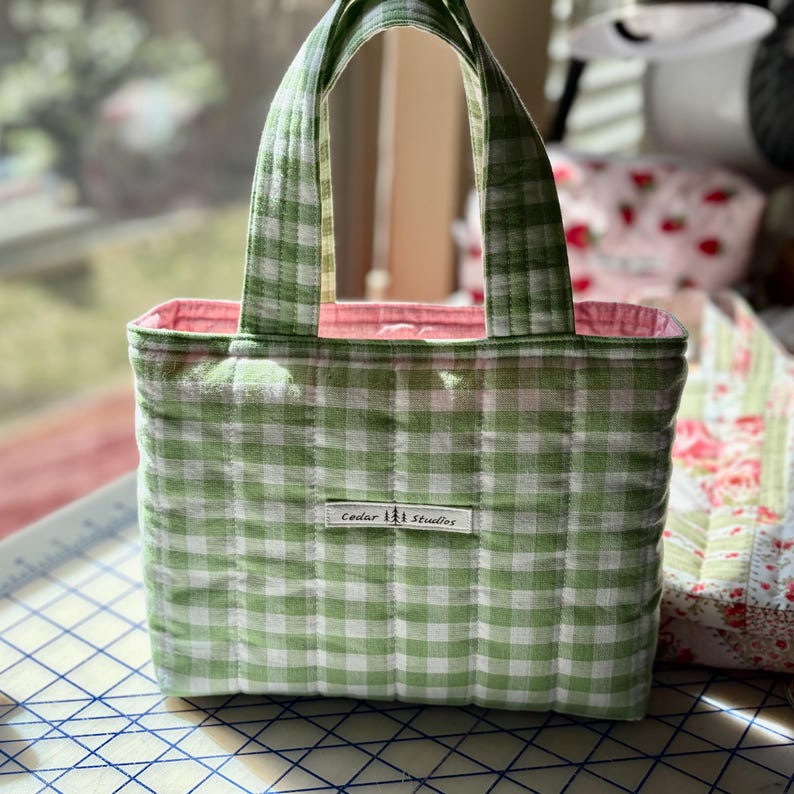 May include: A small, quilted tote bag with a green and white gingham pattern. The bag has a pink interior and matching green handles. A small white tag with the text "Cedar Studios" is sewn on the front.