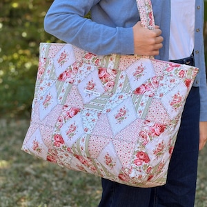 Puede incluir: Un bolso tote acolchado grande con un diseño de patchwork. El bolso presenta una variedad de patrones florales en tonos de rosa, rojo y verde sobre un fondo blanco. El bolso tiene una correa a juego y una cremallera rosa.