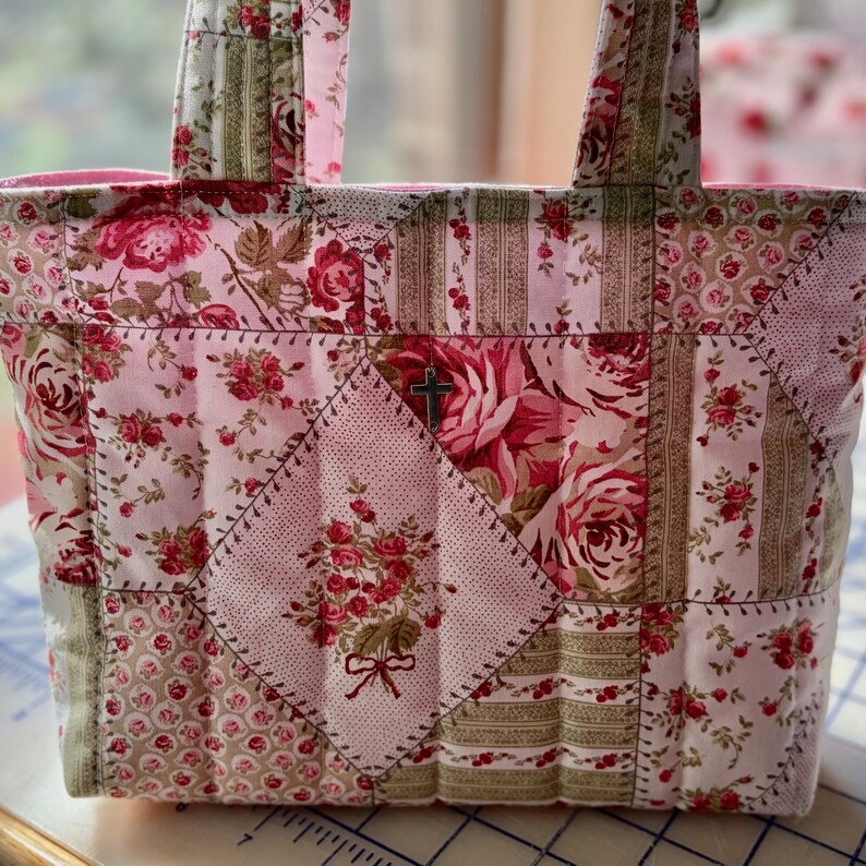 May include: A patchwork tote bag with a floral pattern in shades of pink, red, and green. The bag features a cross emblem and is constructed with various fabric pieces, showcasing a quilted design. The handles are made of the same patterned fabric.