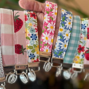 May include: A collection of fabric wrist keychains with various patterns, including strawberries, daisies, and gingham. The keychains feature silver-toned hardware and a variety of colors, such as pink, blue, and white. The keychains are held in a hand.