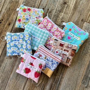 Puede incluir: Una colección de pequeñas bolsas con cremallera con varios diseños florales, frutales y temáticos de café. Las bolsas presentan una variedad de colores, incluyendo azul, rosa y verde, e incluyen el texto "Cedar Studios". Cada bolsa tiene un pequeño anillo de metal.