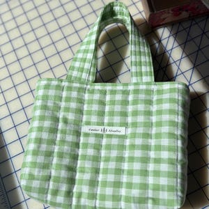 May include: A small, quilted tote bag with a green and white gingham pattern. The bag has two handles and a rectangular label that reads "Cedar Studios". The bag is made of a soft, padded fabric.