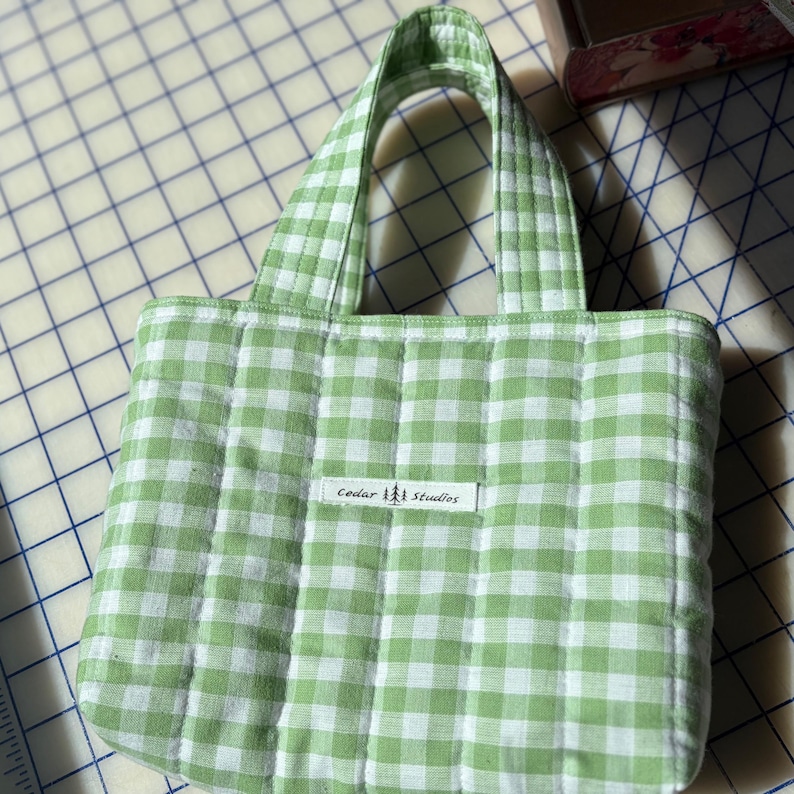 May include: A small, quilted tote bag with a green and white gingham pattern. The bag has two handles and a rectangular label that reads "Cedar Studios". The bag is made of a soft, padded fabric.