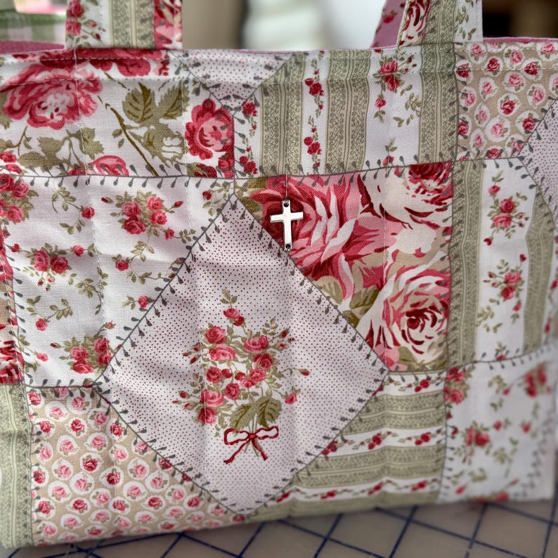 May include: A patchwork tote bag with a floral pattern in shades of pink, white, and green. The bag features a silver cross charm and is constructed with various fabric squares. The design includes embroidered details and a red bow.