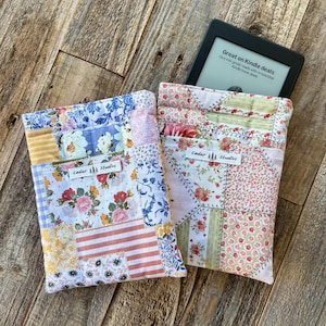 May include: Two patchwork fabric Kindle e-reader sleeves with floral and geometric patterns. One sleeve has a blue, white, and pink floral design, and the other has a pink and green floral design. The text "Cedar Studios" is on both sleeves.