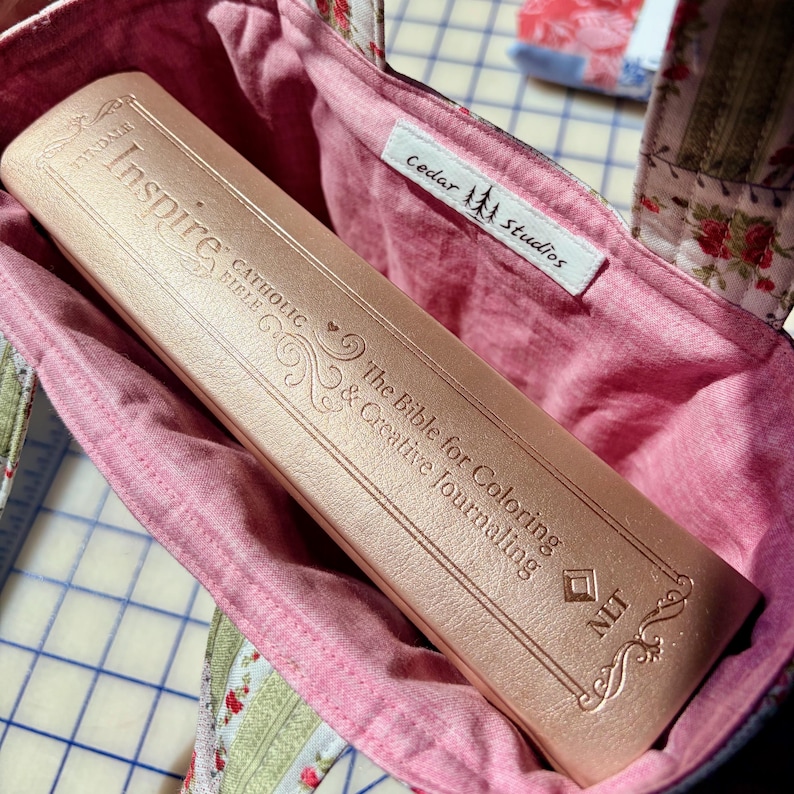 May include: A rose gold Catholic Bible titled "Inspire" for coloring and creative journaling, inside a pink fabric bag. The bag has a floral pattern and a tag that says "Cedar Studios."