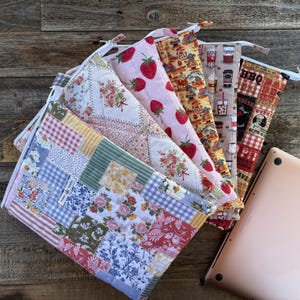 Puede incluir: Una colección de fundas acolchadas para portátiles con varios diseños, incluyendo fresas, motivos florales y un tema de barbacoa. Cada funda tiene cierre de cremallera y un cordón blanco. Un portátil rosa dorado es parcialmente visible.