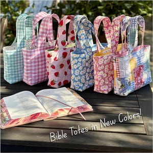 May include: A collection of six quilted fabric tote bags in various patterns including light blue gingham, pink gingham, strawberry print, daisy print, and floral patchwork, arranged behind an open book with the text Bible Totes in New Colors.