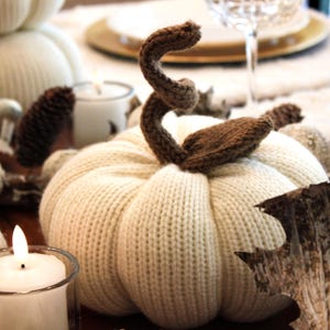 May include: A white knitted pumpkin with a brown knitted stem and leaf. The pumpkin is sitting on a wooden table with a candle and other fall decorations.