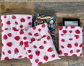 Könnte beinhalten: Rosa Stoffbuch- und E-Reader-Hüllen mit einem sich wiederholenden Muster aus roten Erdbeeren und grünen Blättern. Ein Buch und ein E-Reader befinden sich in den Hüllen. Die Hüllen liegen auf einer Holzoberfläche.