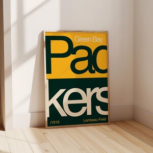May include: A framed poster with a green and yellow colour scheme. The poster features the text "Green Bay Packers" in a bold, retro font. The text "Lambeau Field" and "1919" are also included on the poster.