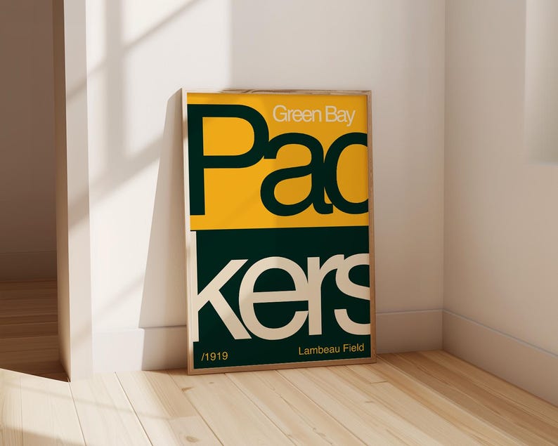 May include: A framed poster with a green and yellow colour scheme. The poster features the text "Green Bay Packers" in a bold, retro font. The text "Lambeau Field" and "1919" are also included on the poster.