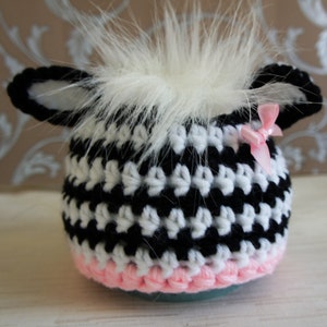 May include: A black and white crocheted hat with a pink trim and a white fur pom-pom on top. The hat has a small pink bow on the side.
