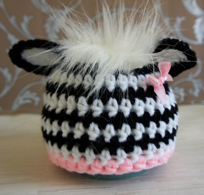 May include: A black and white crocheted hat with a pink trim and a white fur pom-pom on top. The hat has a small pink bow on the side.
