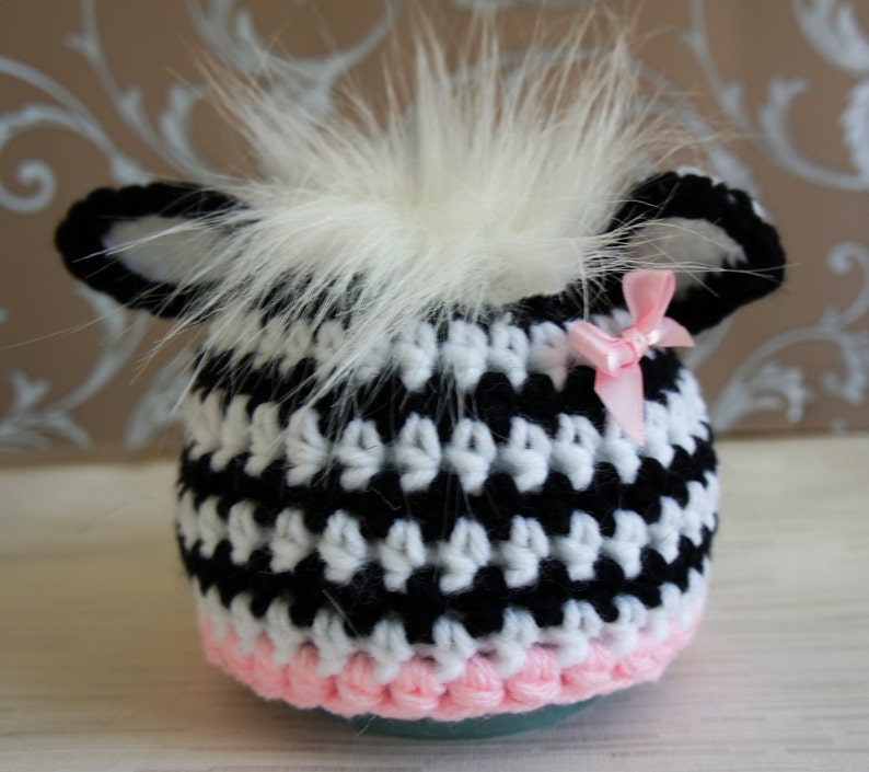 May include: A black and white crocheted hat with a pink bow and a white fur pom-pom. The hat has a pink crocheted brim.
