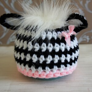 May include: Black and white crocheted hat with a pink trim and a white fur pom-pom on top. The hat has two small ears and a pink bow.
