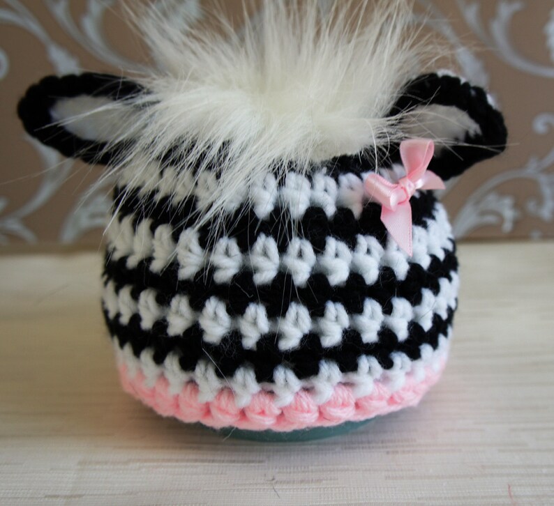 May include: Black and white crocheted hat with a pink trim and a white fur pom-pom on top. The hat has two small ears and a pink bow.