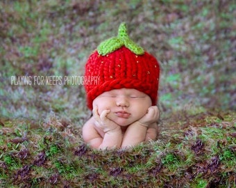 May include: A baby wearing a red knitted strawberry hat with green leaves. The baby is lying on a bed of moss.