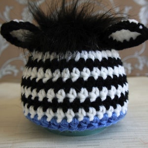 May include: Black and white crocheted hat with blue trim and a black fur pom-pom on top. The hat has two white crocheted ears.