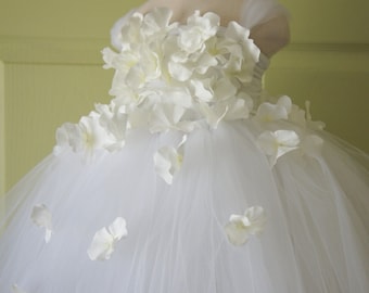 May include: White tulle tutu dress with a white flower embellishment around the bodice. The dress is on a white dress form.