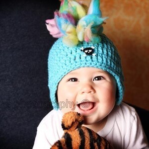 May include: A baby wearing a blue crocheted hat with a colorful pom-pom on top. The hat has a black button on the front. The baby is smiling and holding a brown and black striped stuffed animal.