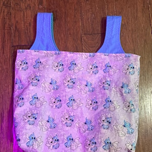 Puede incluir: Un bolso tote con un patr&oacute;n repetido del personaje Stitch en azul y blanco sobre un fondo lavanda. El bolso tiene correas azules y un interior azul.