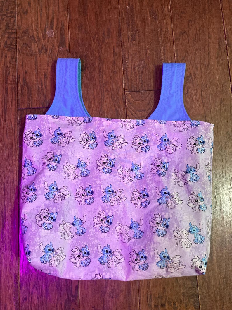 Puede incluir: Un bolso tote con un patr&oacute;n repetido del personaje Stitch en azul y blanco sobre un fondo lavanda. El bolso tiene correas azules y un interior azul.