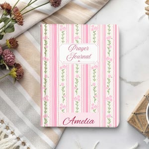 May include: A pink and white striped prayer journal with floral accents and the name "Amelia" at the bottom. The cover features the words "Prayer Journal" in an elegant script. The journal is set on a striped cloth with flowers.