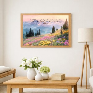 May include: A rectangular wall art print in a light wood frame featuring a landscape painting of a mountain range, evergreen trees, and a meadow of yellow and purple wildflowers. The text reads: The LORD is my strength and my shield; my heart trusts in him, and he helps me. My heart leaps for joy, and with my song I praise him. PSALM 28:7.