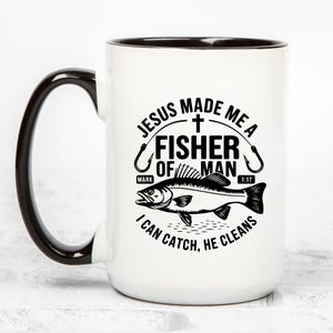 May include: White ceramic mug with a black interior and handle. The mug features a black and white graphic of a fish and the text "JESUS MADE ME A FISHER OF MAN I CAN CATCH, HE CLEANS". The text also includes the bible verse Mark 1:17.