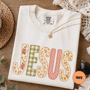 May include: An ivory t-shirt featuring the word JESUS in large, block-style letters. Each letter has a unique pattern: floral prints with roses and small yellow flowers, a green and white gingham check, and a pink polka dot design.
