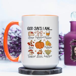 May include: White ceramic mug with an orange handle and rim. The mug features the text "GOD SAYS I AM..." and illustrations of a cake, bow, coffee cup, berries, pumpkin, leaves, mushrooms, acorns, and a plant. The mug is on a dark grey coaster.