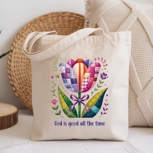 May include: A cream tote bag with a vibrant floral design. The flower is composed of patchwork-style petals in shades of purple, pink, and orange, with green leaves and a purple bow. The text "God is good all the time" is printed below.
