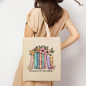 May include: Beige tote bag featuring a colorful floral design and the text "Women Of The Bible." The design includes stacked books with names like "Loyal," "Brave," and "Patience." The bag is held by a person wearing a beige dress.
