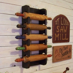 Peut inclure: Un porte-rouleaux en bois noir avec cinq rouleaux &agrave; p&acirc;tisserie en bois. Les rouleaux &agrave; p&acirc;tisserie ont des poign&eacute;es de diff&eacute;rentes couleurs. Le porte-rouleaux est mont&eacute; sur un mur en bois blanc. Il y a deux panneaux sur le mur, l'un qui dit "St. Louis Saw Mill" et l'autre qui dit "Old Paint Saddle Co. Tested and True since 1842."