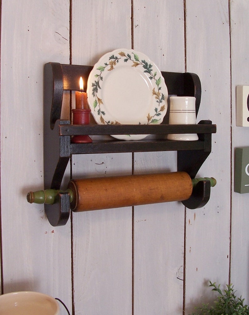 May include: A dark brown wooden wall shelf featuring a top ledge holding a white plate with a green and brown leaf pattern, a red candle, and a white cup. Below the shelf, a wooden rolling pin with green handles rests in a built-in holder.