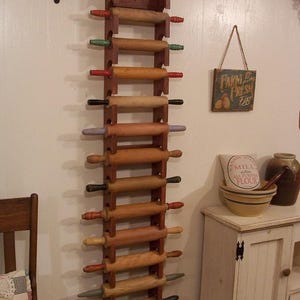 Op de afbeelding: Een houten deegrollerrek met meerdere deegrollers. Het rek is aan een witte muur bevestigd. De deegrollers hebben houten cilinders en kleurrijke handvatten in rood, groen, zwart en andere kleuren. Een bord met "Farm Fresh Eggs" hangt aan de muur.