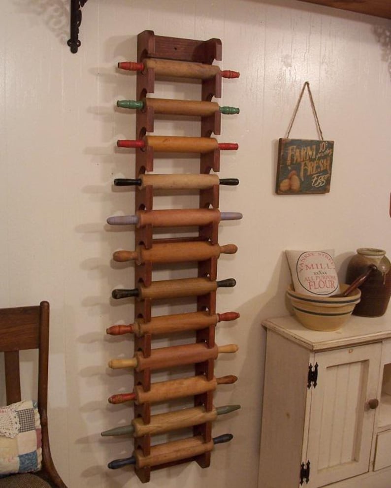 Op de afbeelding: Een houten deegrollerrek met meerdere deegrollers. Het rek is aan een witte muur bevestigd. De deegrollers hebben houten cilinders en kleurrijke handvatten in rood, groen, zwart en andere kleuren. Een bord met "Farm Fresh Eggs" hangt aan de muur.
