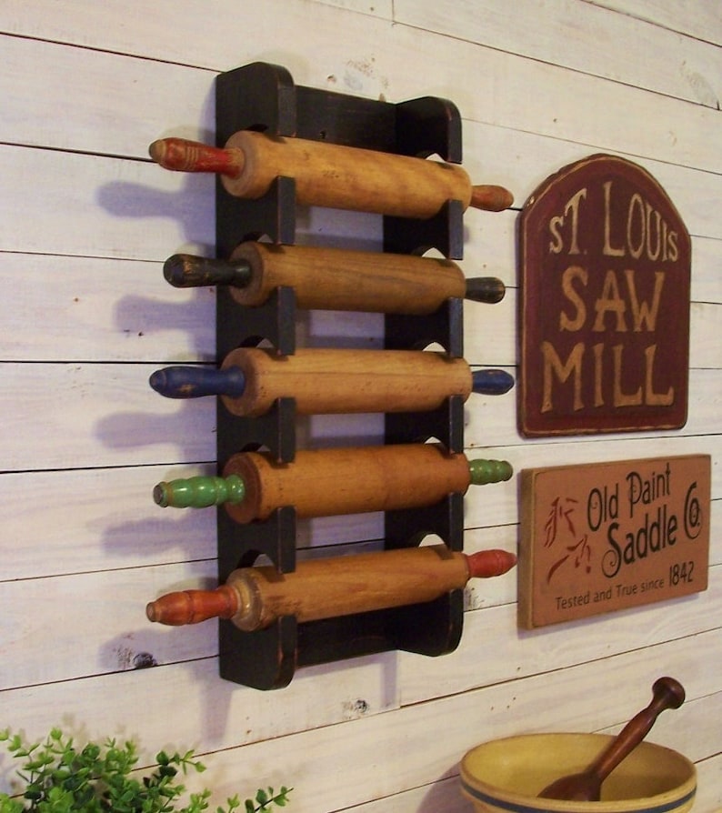 Peut inclure: Un porte-rouleaux en bois noir avec cinq rouleaux &agrave; p&acirc;tisserie en bois. Les rouleaux &agrave; p&acirc;tisserie ont des poign&eacute;es de diff&eacute;rentes couleurs. Le porte-rouleaux est mont&eacute; sur un mur en bois blanc. Il y a deux panneaux sur le mur, l'un qui dit "St. Louis Saw Mill" et l'autre qui dit "Old Paint Saddle Co. Tested and True since 1842."