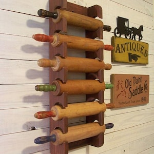 Op de afbeelding: Een houten deegrollerrek met zes vintage deegrollers. Het rek is donkerbruin en de deegrollers hebben handvatten in verschillende kleuren. Een bord met de tekst "Antiek" en een ander bord met de tekst "Old Paint Saddle Co" zijn ook zichtbaar.