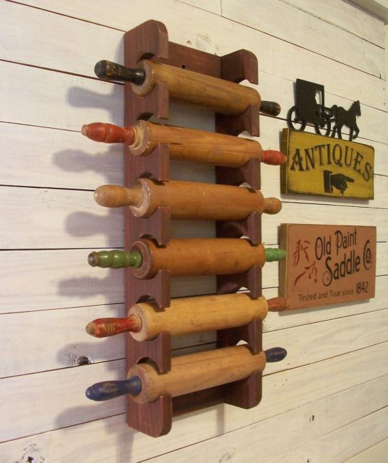 Op de afbeelding: Een houten deegrollerrek met zes vintage deegrollers. Het rek is donkerbruin en de deegrollers hebben handvatten in verschillende kleuren. Een bord met de tekst "Antiek" en een ander bord met de tekst "Old Paint Saddle Co" zijn ook zichtbaar.