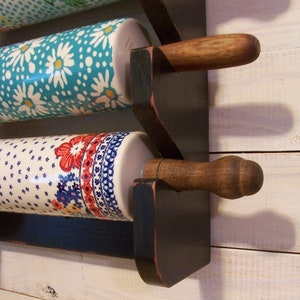 May include: A black wooden rolling pin rack with three ceramic rolling pins. The rolling pins have colorful floral patterns. The rack is mounted on a white wall.