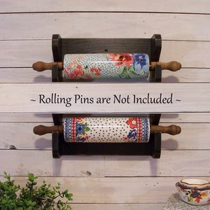 May include: A black wooden rolling pin rack with two shelves, each holding a colorful ceramic rolling pin. The rack is mounted on a white wood wall. The text "~ Rolling Pins are Not Included ~" is written on a white wooden sign above the rack.