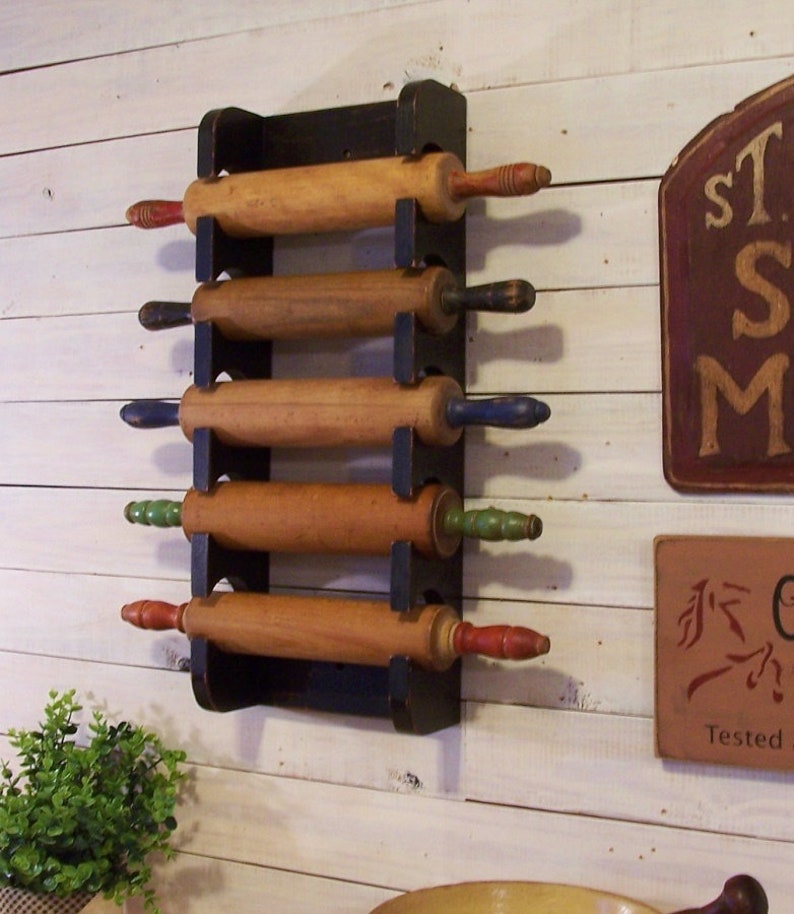 Peut inclure: Un porte-rouleaux en bois noir avec cinq rouleaux &agrave; p&acirc;tisserie en bois. Les rouleaux &agrave; p&acirc;tisserie ont des poign&eacute;es de diff&eacute;rentes couleurs, notamment rouge, bleu et vert. Le support est mont&eacute; sur un mur en bois blanc.