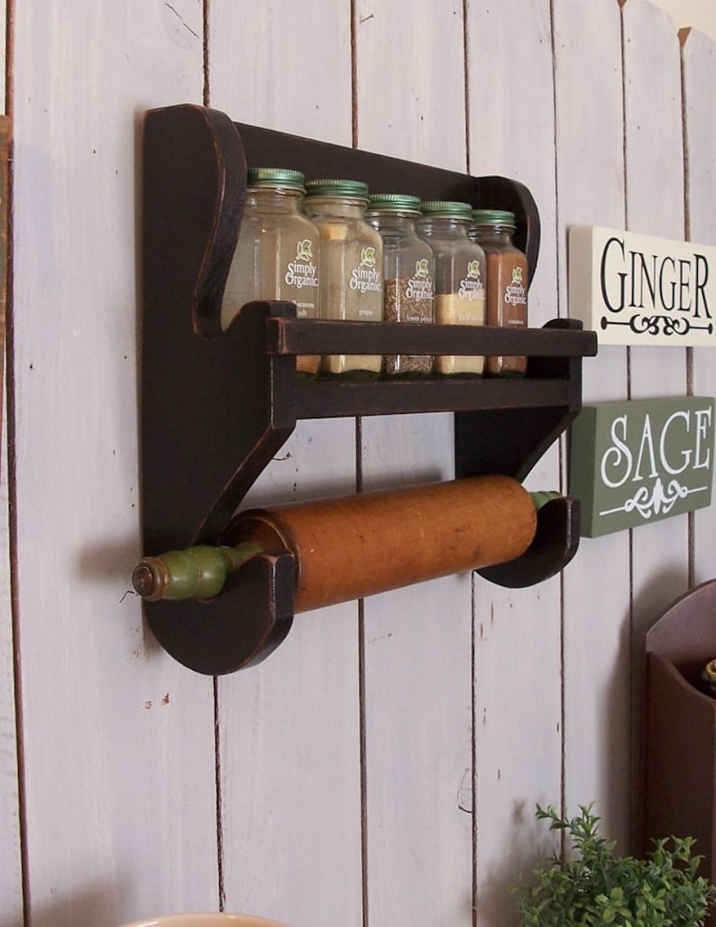 May include: A dark brown wooden wall-mounted spice rack featuring a top shelf holding five glass spice jars with green lids and a lower rod supporting a wooden rolling pin with green handles. The rack is mounted on a white vertical plank wall.