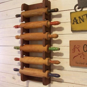 Op de afbeelding: Een houten deegrollerrek met zes deegrollers. De deegrollers hebben gekleurde handvatten in rood, groen, blauw en zwart. Het rek is donkerbruin en is gemonteerd op een witte houten muur.